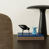 Black bird figurine on blue book stacked with a textured beige book, alongside a black lamp. Elegant contemporary decor.