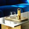 "Gold bottle opener and clear glass on a wooden tray atop a marble table, with blue sofa in the background."
