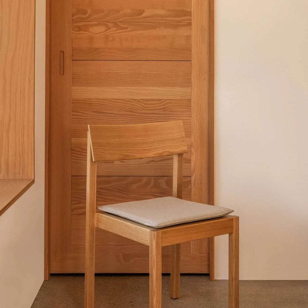 Natural wood chair with a smooth finish and a light gray cushion against a warm wood backdrop. Simple and modern design.