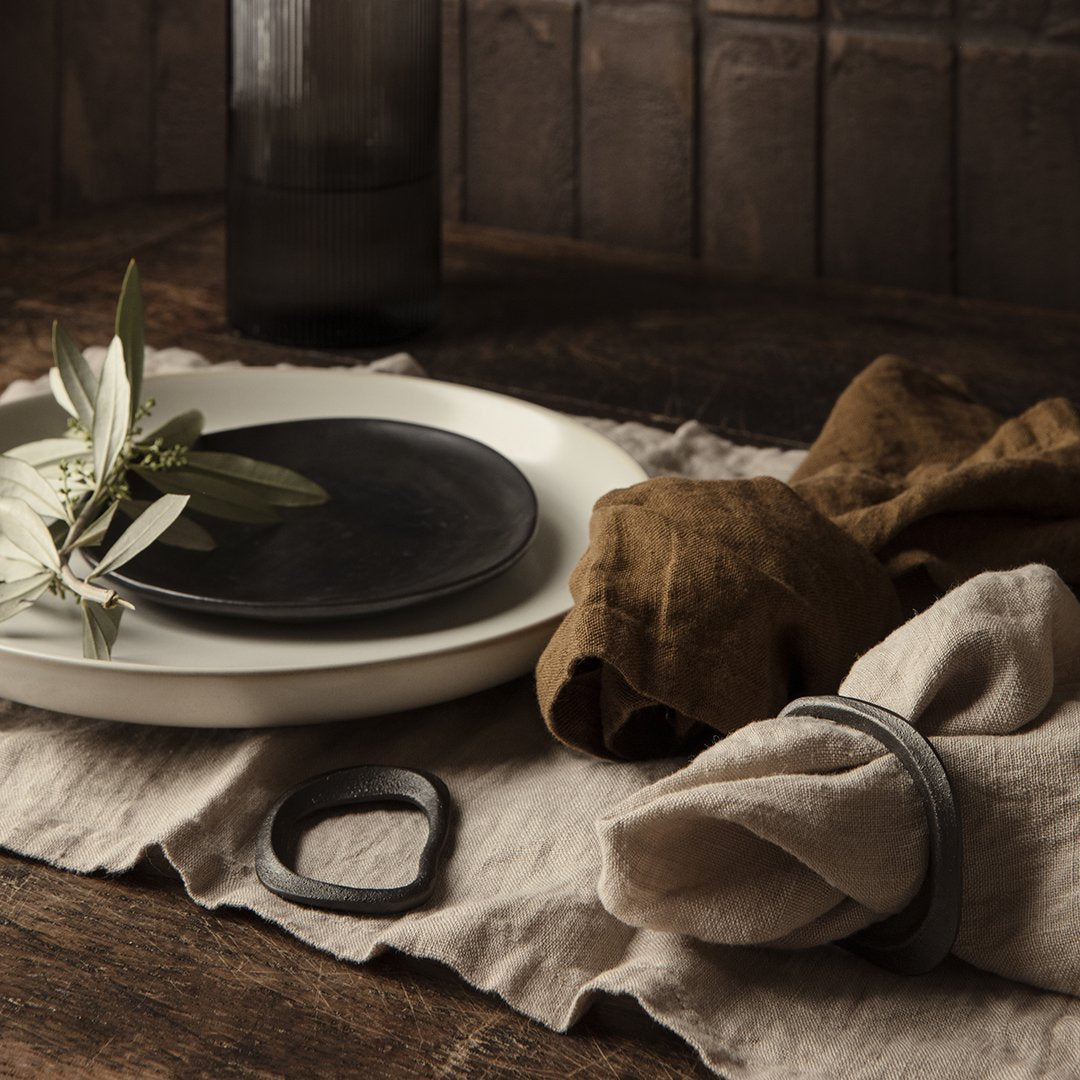 "Neutral-toned tableware with black plates, earthy linen napkins, and a simple green sprig, styled on a textured beige cloth."