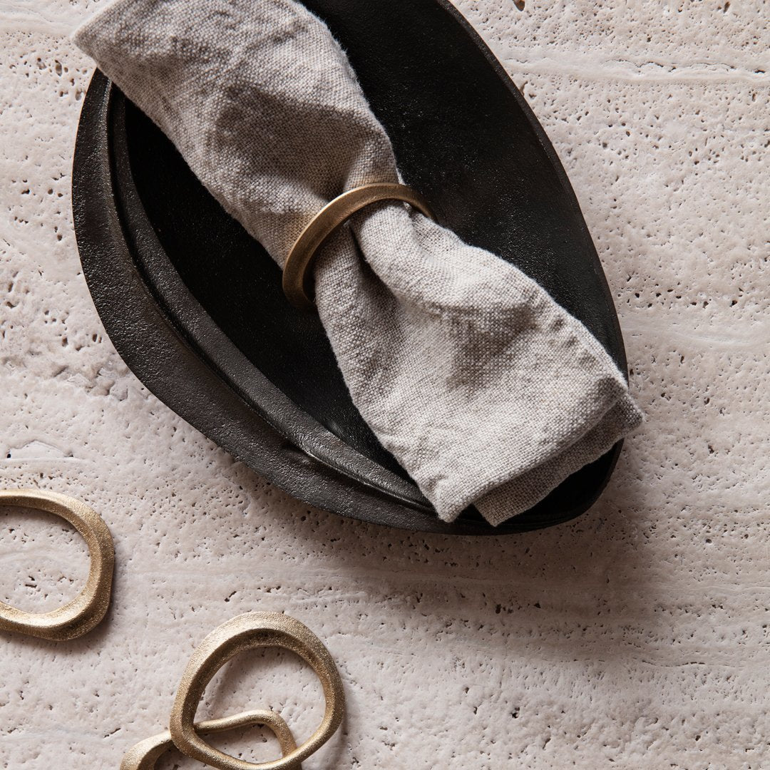 Linen napkin wrapped in a gold ring on a textured black ceramic plate; neutral tones and elegant shapes.