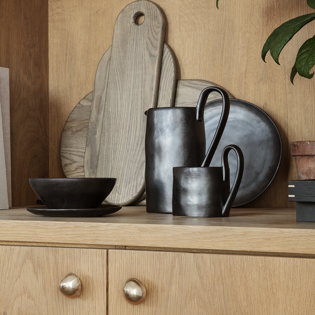 Matte black pottery set with a smooth, minimalist finish, displayed against natural wood and green foliage.