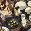 Rustic table setting with earthy tones, green ceramics, and a brown patterned cloth featuring vibrant fruits and glassware.