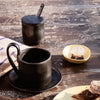 Black ceramic cup and saucer set with a matte finish, paired with a textured black sugar jar and a pastel yellow dish.
