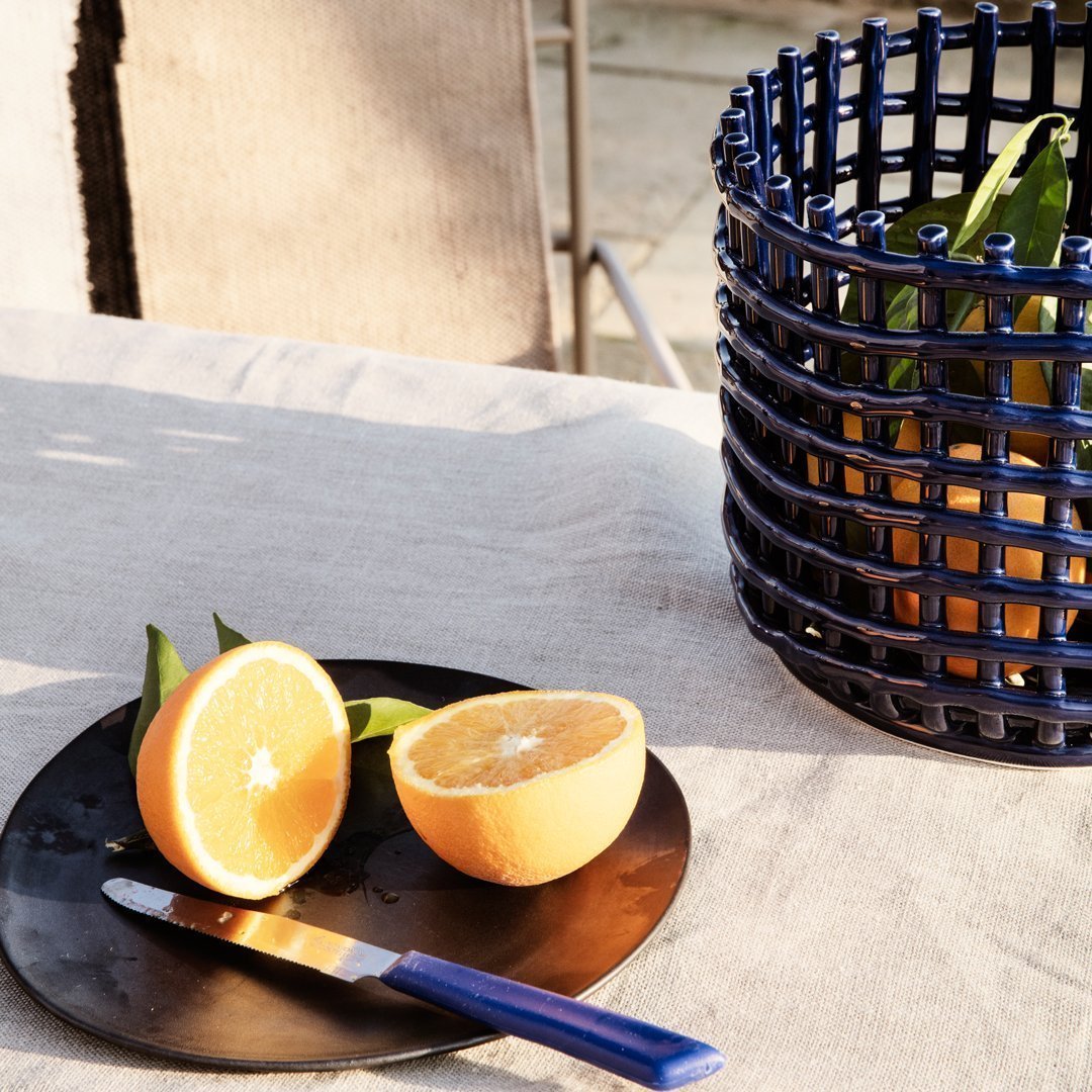 Blue braided basket with oranges on a black plate and silver knife on a light gray tablecloth. Fresh, vibrant colors.