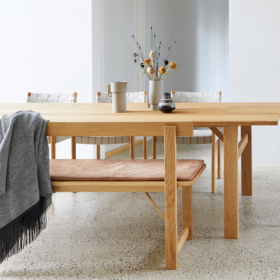 Wooden dining table with a natural finish, beige bench cushion, and grey throw, complemented by soft textured patterns.