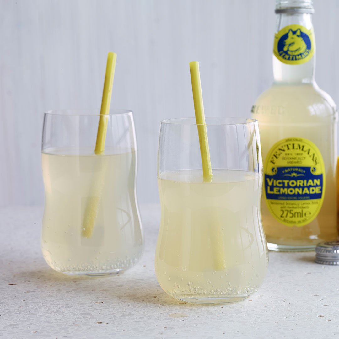 Two clear glasses of light yellow lemonade with yellow straws, set beside a bottle, against a neutral background.
