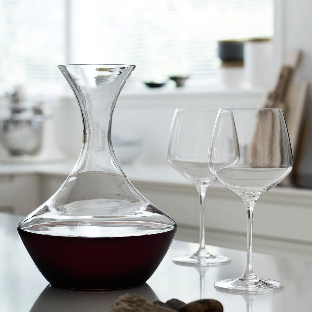 Elegant glass decanter with a sleek design, featuring a clear body and rich dark red wine at the base. Two clear wine glasses.