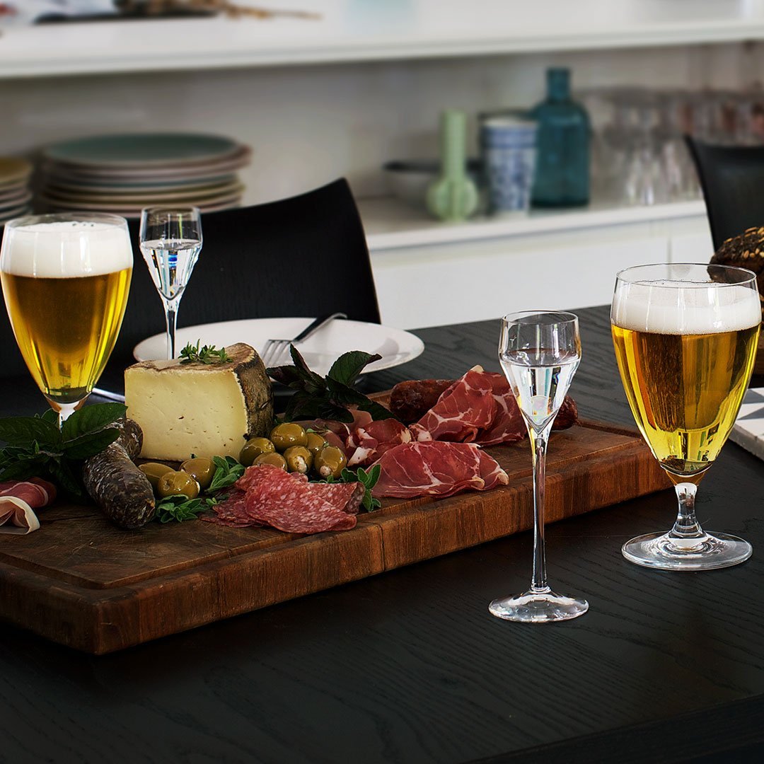 "Wooden platter with dark green leaves, assorted cheeses and meats, paired with golden beers and clear glasses."