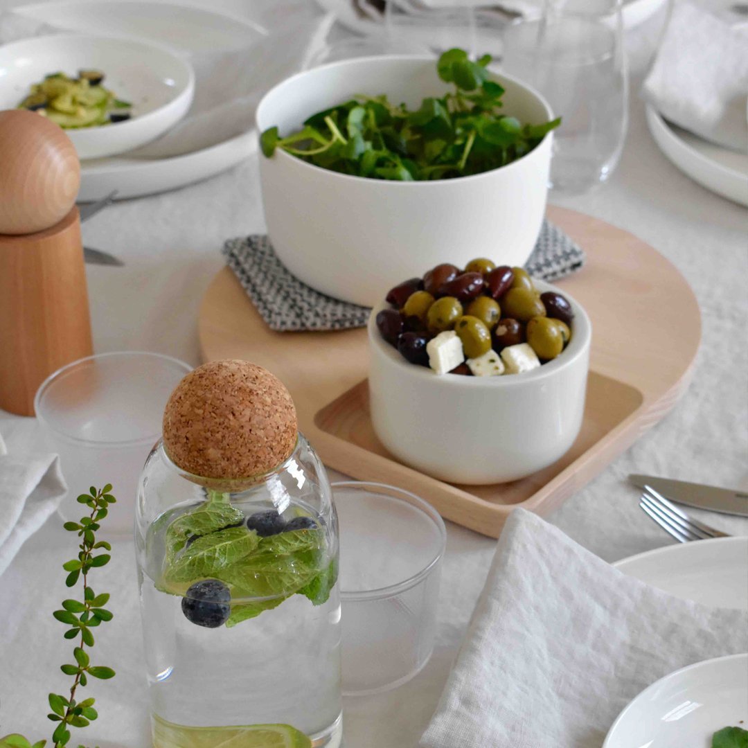 "White bowls with greens and olives on a light wooden tray, set against a neutral tablecloth."