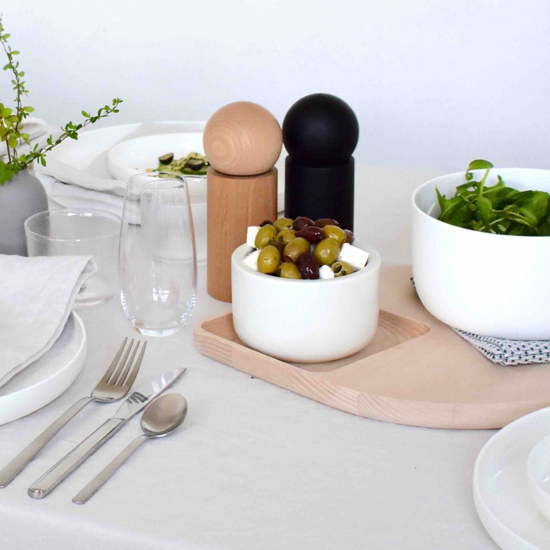 White bowl with olives, black and beige salt shakers, green salad in a bowl, neutral table setting with subtle patterns.