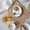 "White plates on a textured wooden tray, featuring a beige cookie, gold drink, and a soft cream-colored towel."