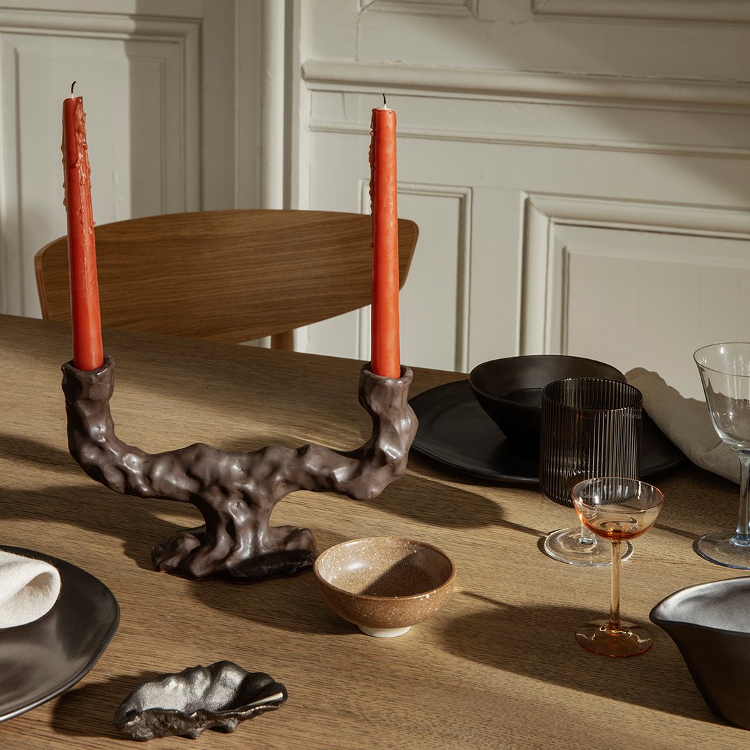 Brown sculptural candleholder with orange candles, set on a wooden table with black, cream, and gold dinnerware.