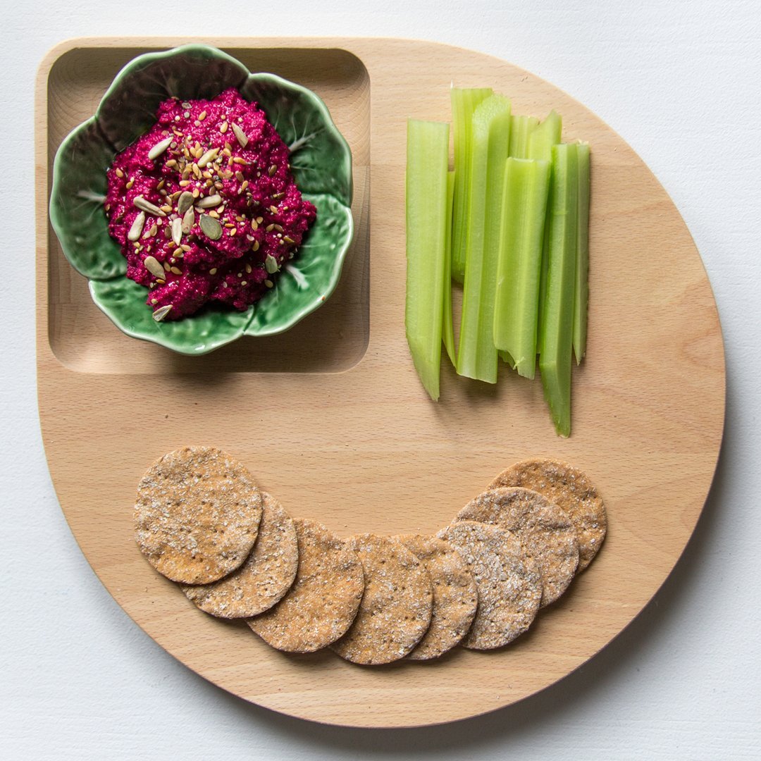 Colorful wooden platter with green bowl, vibrant pink dip, fresh celery sticks, and golden-brown crackers in a playful arrangement.