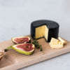 Black cheese wheel, creamy yellow interior, garnished with green figs and brown butter on a wooden board.