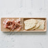 Wooden serving tray with pink prosciutto and light beige crackers on a white marble surface.