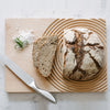 Rustic brown artisan bread with a crusty white pattern, served on a circular wooden board with green garnish and salt.