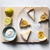 Lemon cheesecake slices arranged on a textured cream-colored board, with vibrant yellow lemons and a teal bowl.