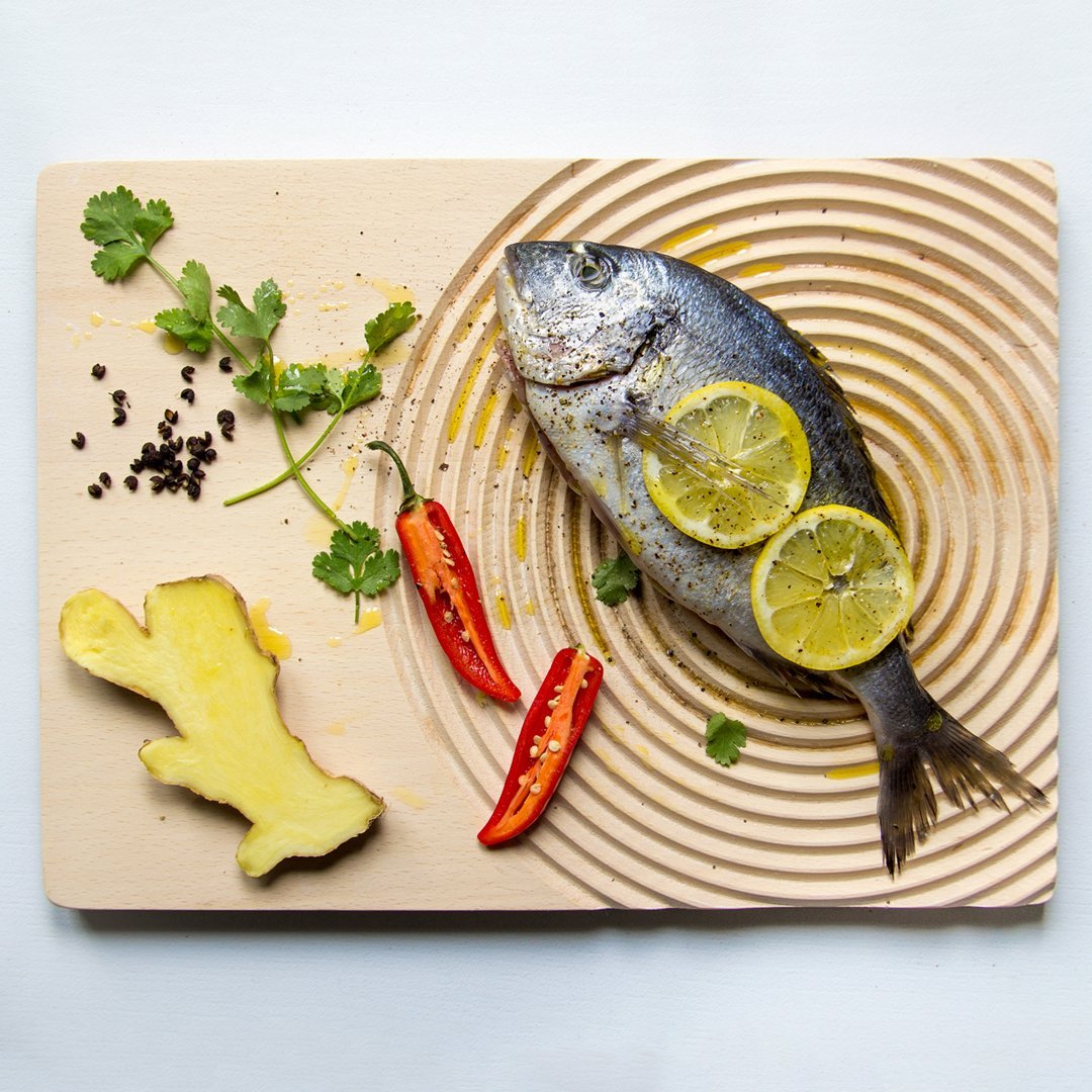 Fresh fish with vibrant yellow lemon slices, red chili, green cilantro, and a textured wooden cutting board.
