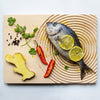 Fresh fish with vibrant yellow lemon slices, red chili, green cilantro, and a textured wooden cutting board.