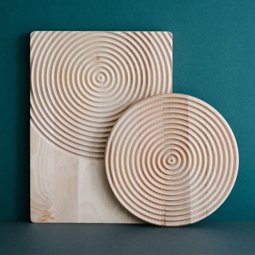 Natural wood serving board and plate with concentric circle patterns, showcasing light beige tones against a teal backdrop.